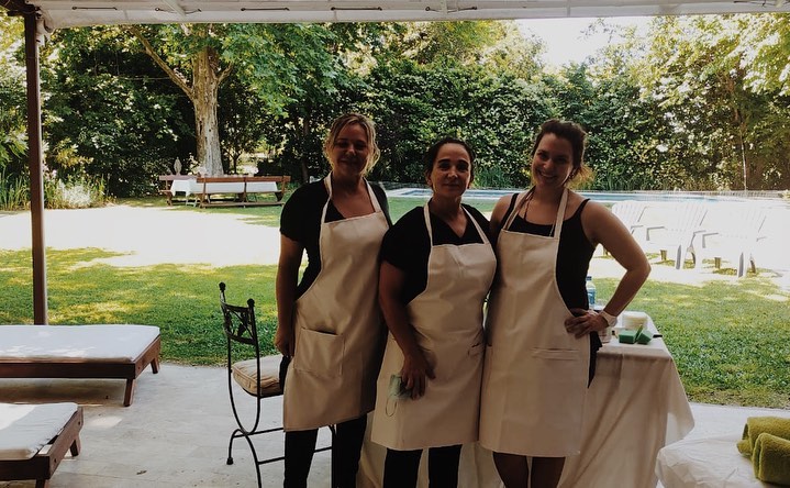 Tres integrantes del staff con delantal, bajo una pérgola, en un evento al aire libre con jardín y pileta